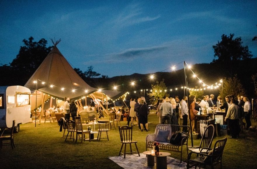 A wedding tipi is set up with indoor and outdoor festoon lighting. People gather inside and out around tables and chairs