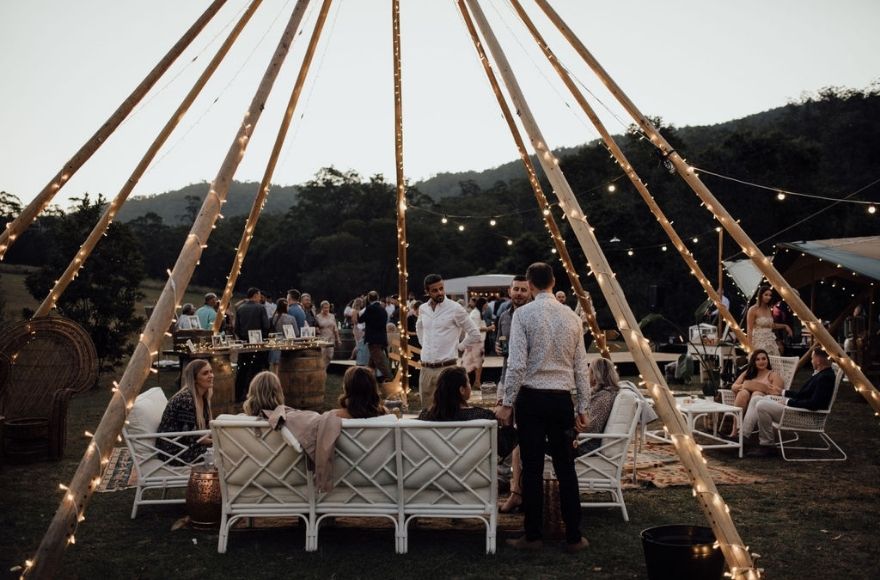 People congregate under an open air tipi frame with fairy lights wrapped around the poles. 