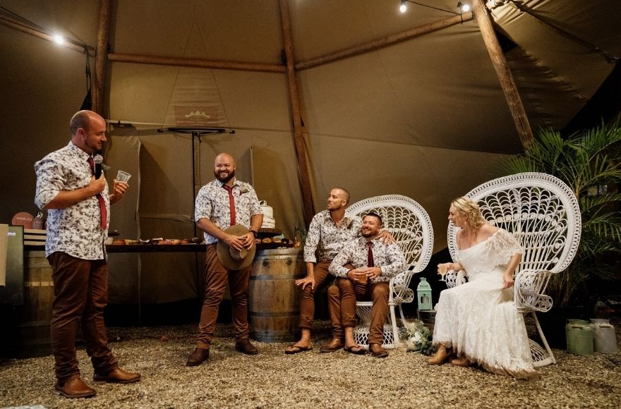 Couple sit on white peacock chairs under a tipi tent. They are talking to two groomsmen standing by a rustic bar cart.