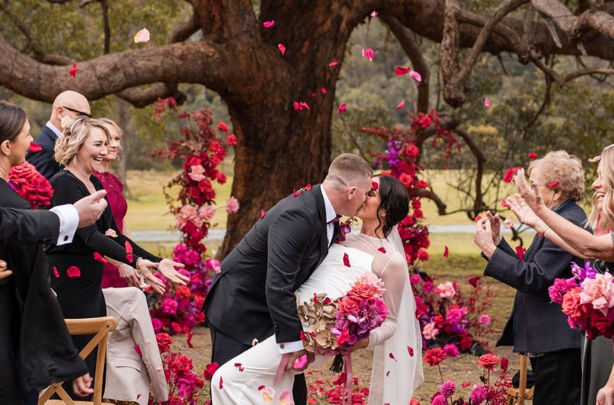 Newly wed couple have their first kiss surrounded by pink, red, and purple floral displays