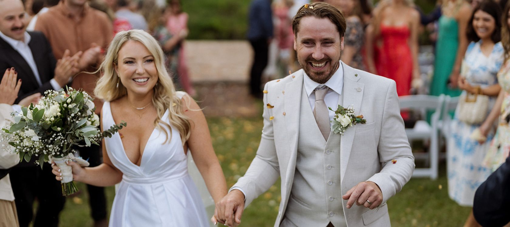 Bride with blonde hair wears a white wedding dress, hold hands with groom wearing taupe 3 piece suit