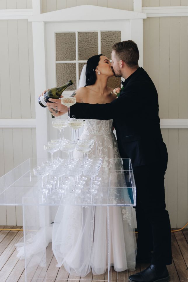 Bride and groom kiss each other while pouring champagne into a tower of coupe glasses.