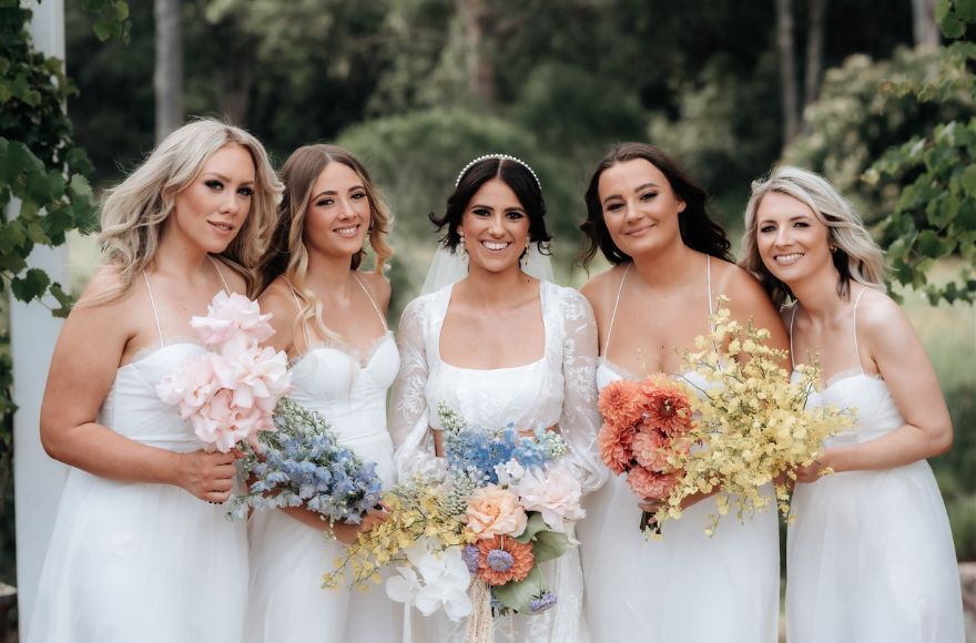 A bride and her bridesmaids all dressed in white carry different bouquets of pastel coloured flowers