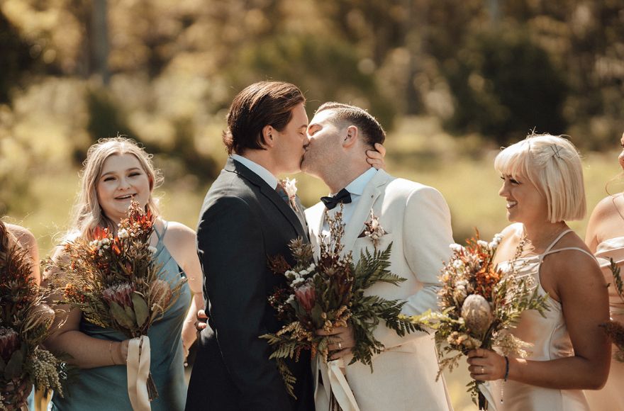 Newly wed grooms share their first kiss with bridesmaids looking on.