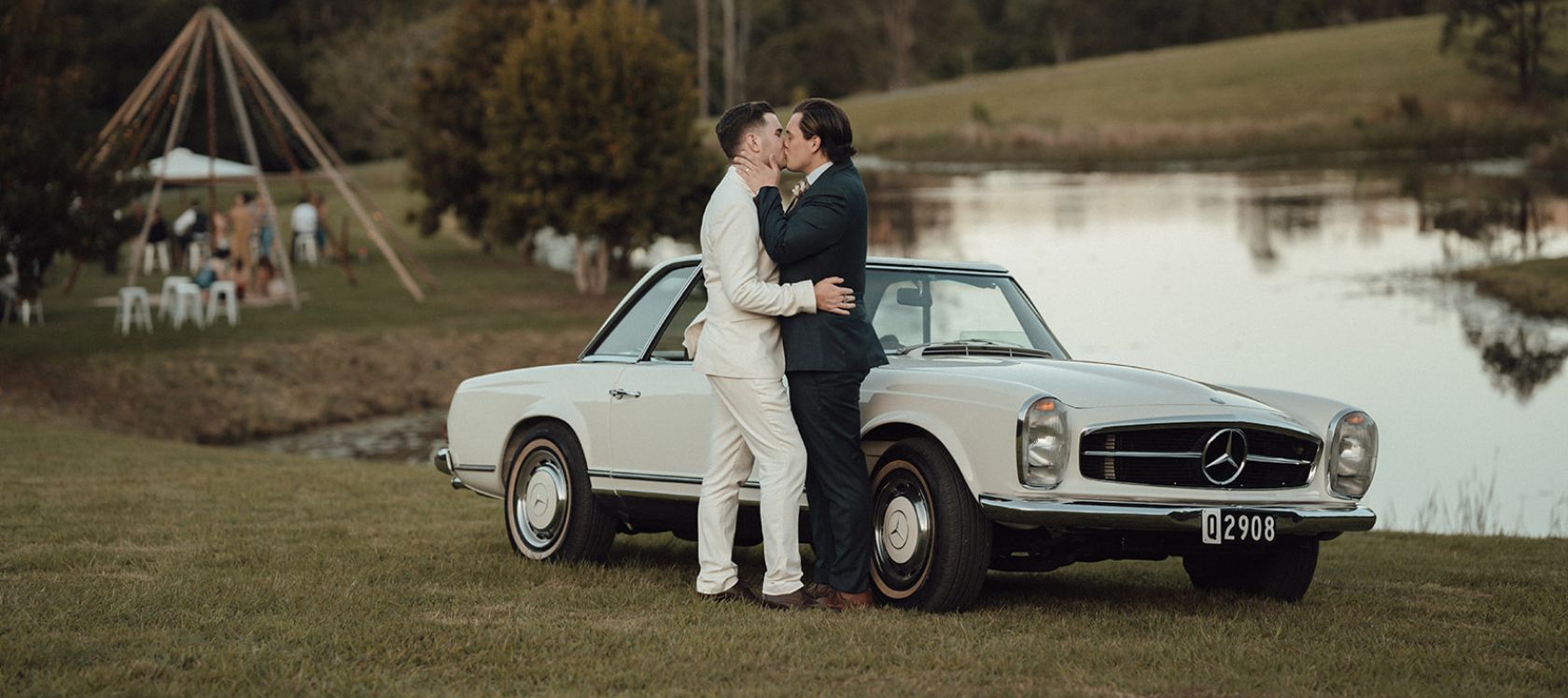 Newly wed grooms kiss by a vintage Mercedes against a lake and forest