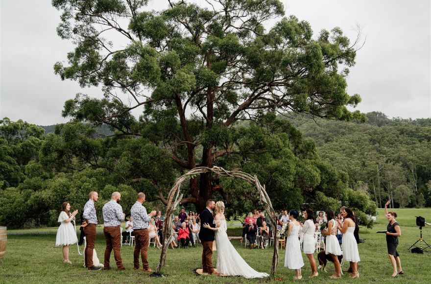 A couple have their wedding ceremony under a rustic arbour, in front of an old tree