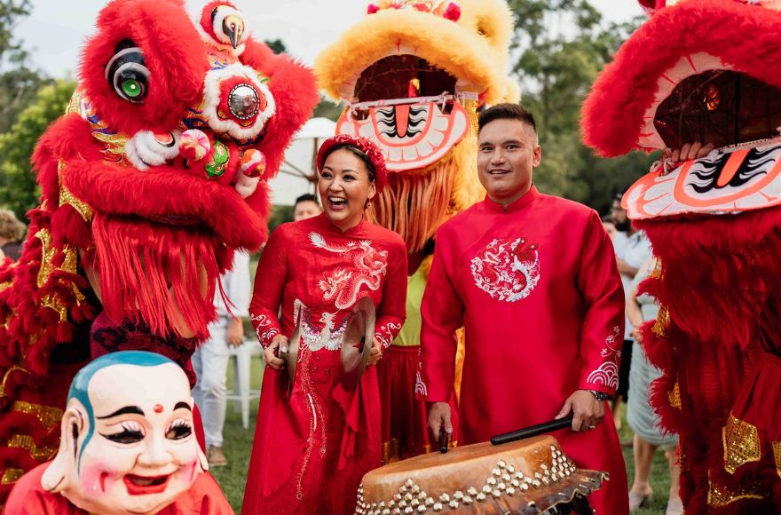 couple wear traditional Vietnamese wedding attire surrounded by dancing dragons and drummers