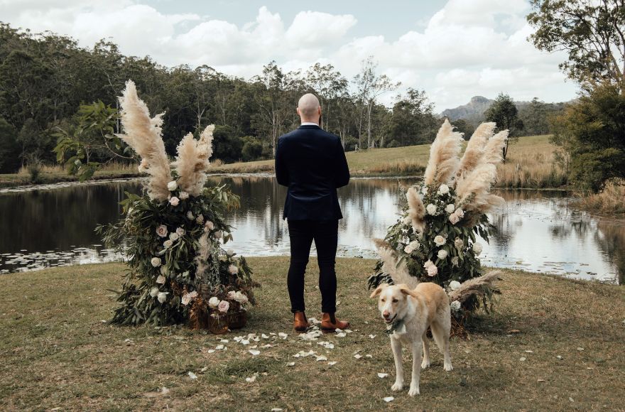 A groom stands at the wedding ceremony location by the lake with his dog