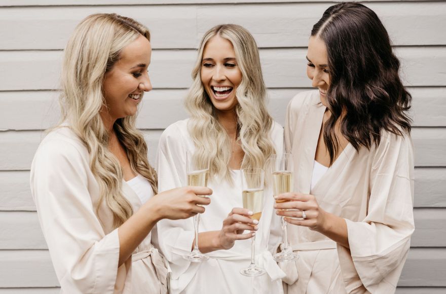 3 women dressed in ivory silk robes toast with champagne flutes