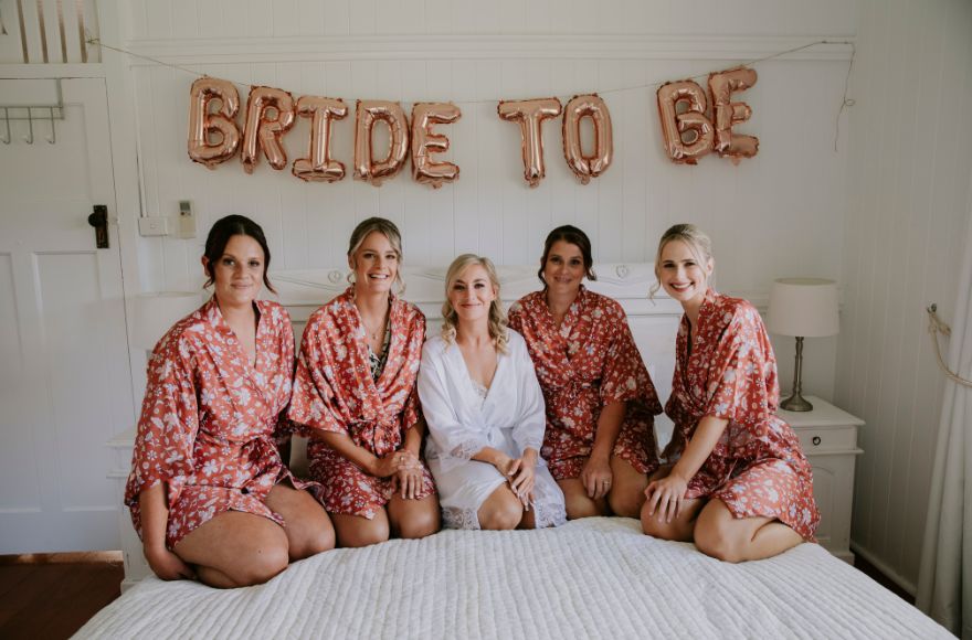 A bride and her 4 bridesmaids sit on the bed together with a sign behind them that  says "Bride To Be"