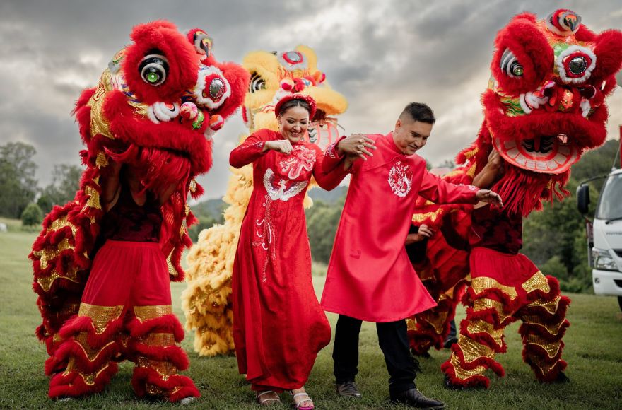 Newlywed couple wear traditional Vietnamese red robes while dancing with dragon dancers on the lawn