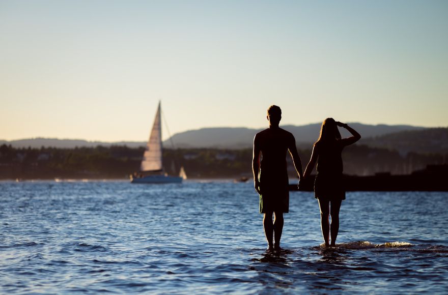 Couple stand in the water holding hands looking out at boats and distant islands