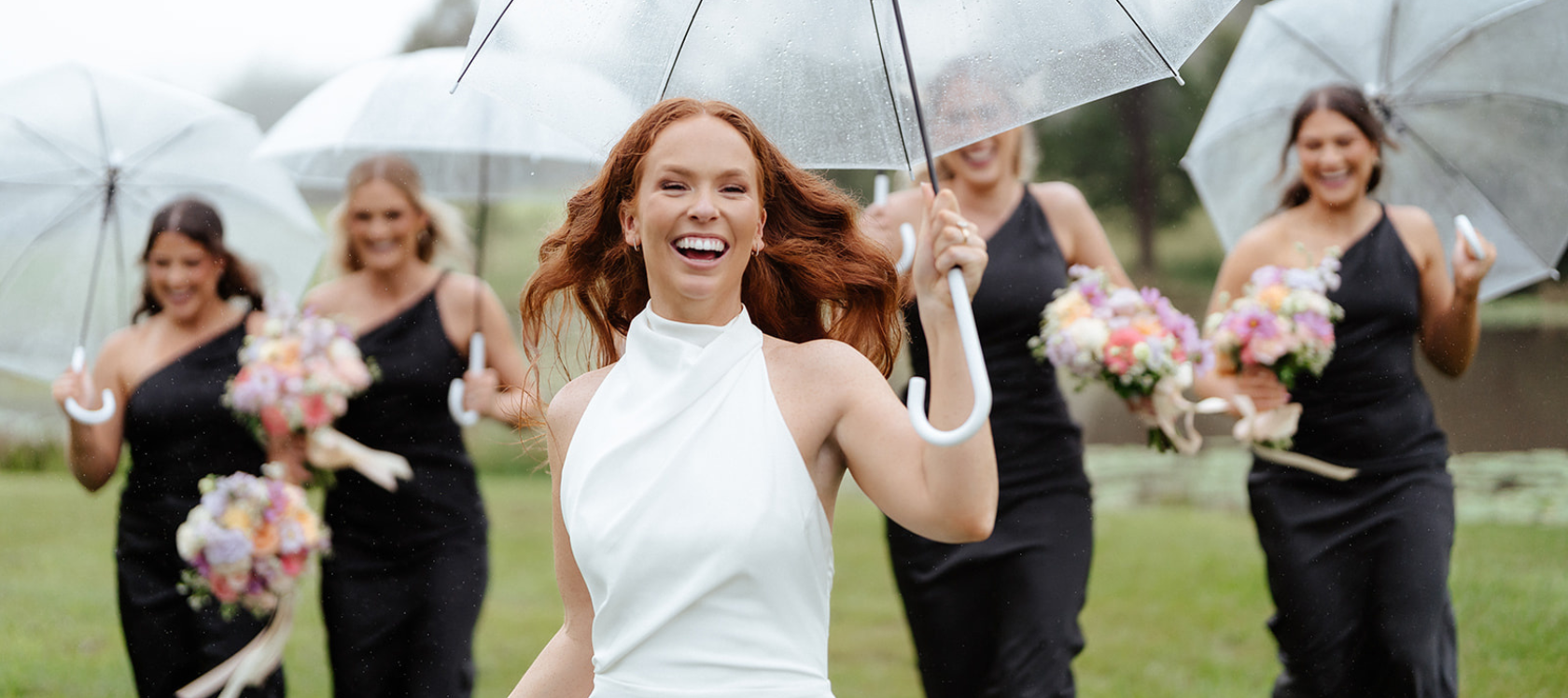 Bride with long red hair poses with bridesmaid in the background. They are holding clear umbrellas in light rain.