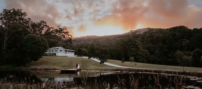View of the Gold Coast Farm House with the lake in the foreground. A couple stands on the jetty with the house in the background. The sun is setting behind a mountain range.