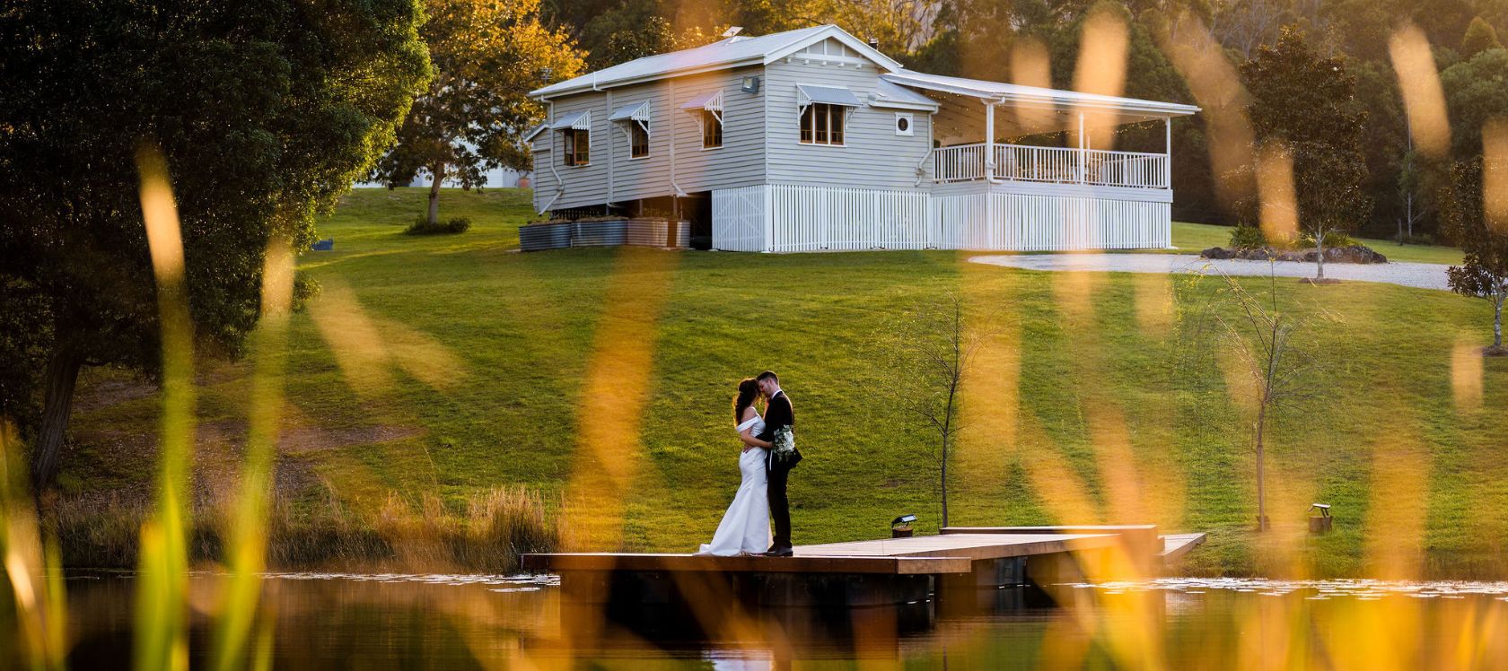Bride and groom stand on a jetty with the Gold Coast Farm House in the background